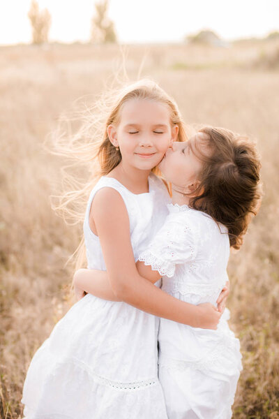 Cute little girls sisters with blond  hair in a summer field at sunset in white dresses with a straw hat