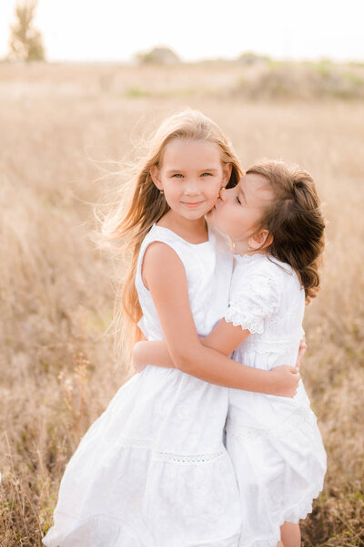 Cute little girls sisters with blond  hair in a summer field at sunset in white dresses with a straw hat