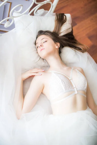 年轻美丽的新娘与黑色长发早晨在家在白色的床上 在白色蕾丝紧身衣和礼服 — 图库照片