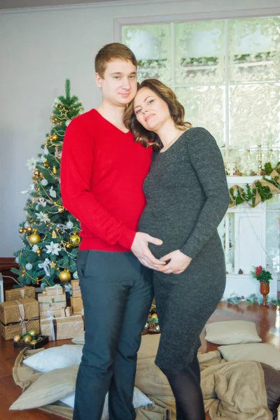 クリスマスツリーを背景に愛する美しい妊娠中のカップルは 新年の前にクリスマス休暇に休息を持っています 大きな妊娠中の腹 — ストック写真