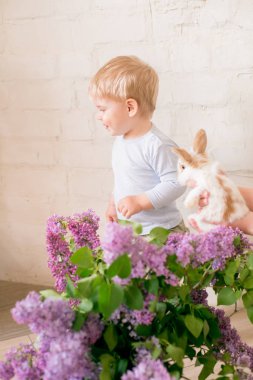 Hafif bir arka plan üzerinde bir hasır sepette leylak çiçekleri ile küçük bunnies ile sarı saçlı küçük sevimli çocuk