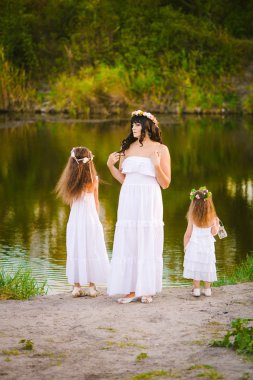 Nehir kıyısında gün batımında bir yaz tarlasında eğlenen beyaz elbiseli kızları ile genç anne