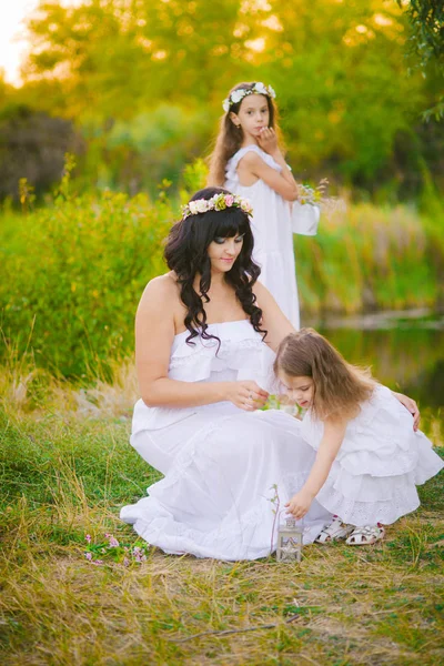 川のそばの夕暮れ時の夏の野原で楽しんでいる白いドレスを着た娘を持つ若い母親 — ストック写真