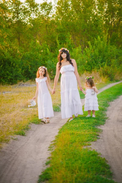 Young mother with her daughters in white dresses having fun in a summer field at sunset by the river