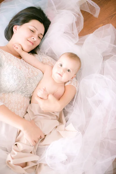 床に横たわっている明るいブドワールの寝室で彼女の腕の中に赤ちゃんを持つ若いママ — ストック写真