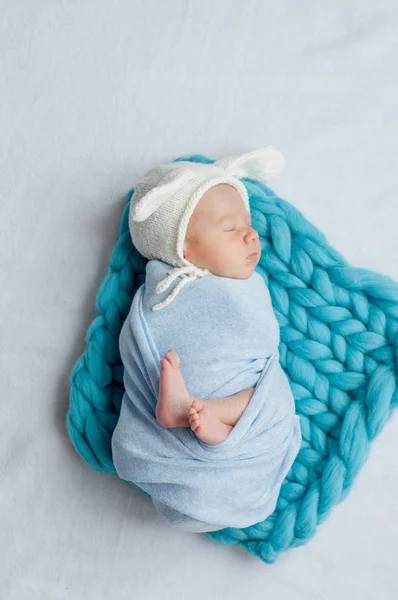 Cute little baby in a blue blanket and a white cap with bunny ears on the bed at home