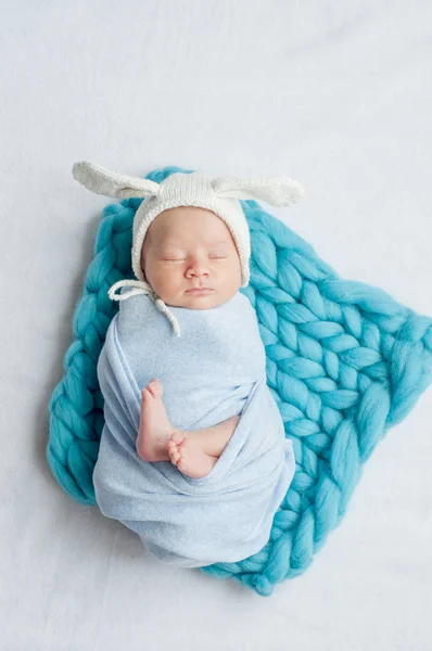 Cute little baby in a blue blanket and a white cap with bunny ears on the bed at home