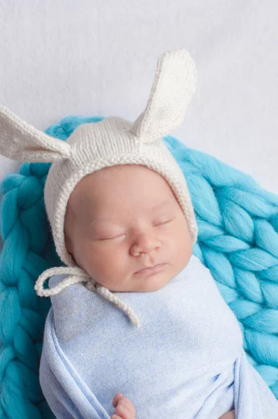 Cute little baby in a blue blanket and a white cap with bunny ears on the bed at home