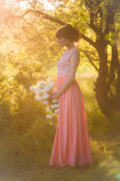 日没時の夏の公園で彼女の手に白い花を持つ長いドレスを着た魅力的な若い女の子 — ストック写真
