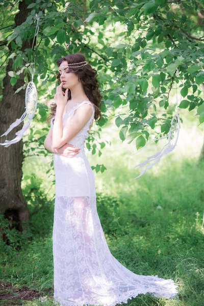 Young attractive girl in a long white dress with a beautiful hairstyle, with white dream catchers in a summer park