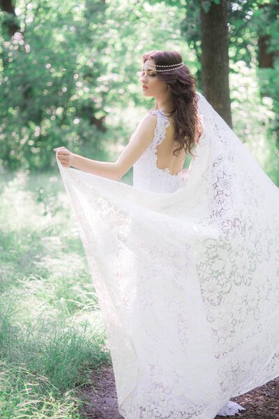 Attractive young girl in a long white dress with a beautiful hairstyle, with a white lace vintage scarf in a summer park