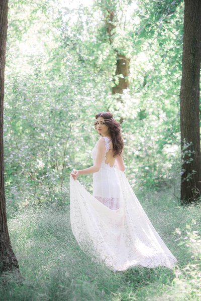 Attractive young girl in a long white dress with a beautiful hairstyle, with a white lace vintage scarf in a summer park