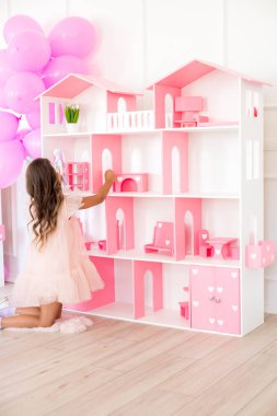 Cute happy girl in a beautiful dress at home plays with a dollhouse and toys clipart