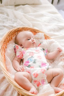 Çiçek baskılı giysiler sevimli bebek evde beyaz bir yatakta bir hasır sepet yatıyor.