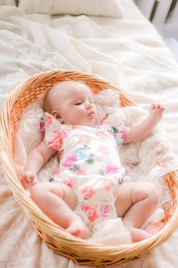 Çiçek baskılı giysiler sevimli bebek evde beyaz bir yatakta bir hasır sepet yatıyor.
