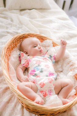 Çiçek baskılı giysiler sevimli bebek evde beyaz bir yatakta bir hasır sepet yatıyor.