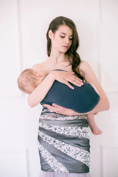 床の上のイブニングドレスの若い美しい母親は明るいスタジオで彼女の腕の中で赤ちゃんを保持します — ストック写真
