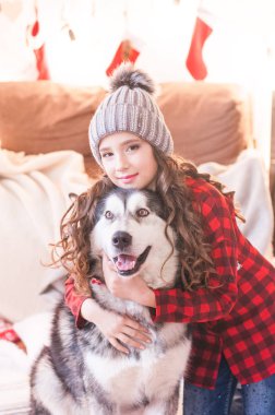 Kırmızı damalı gömlek ve Noel için dekore edilmiş odada evde malamute ile örme şapka sevimli kız. Mutlu noel ruh hali