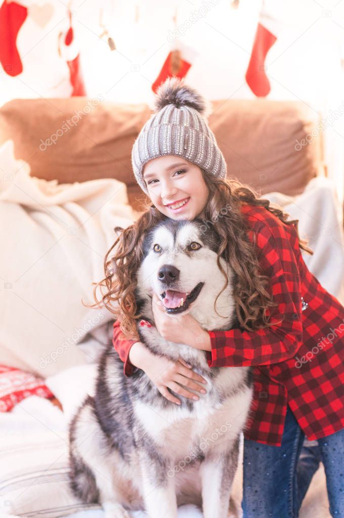 Cute girl in a red checkered shirt and a knitted hat with malamute at home in the room decorated for Christmas. Happy christmas mood