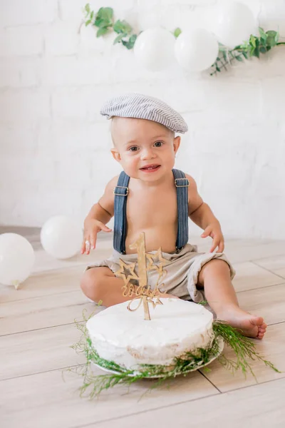 Little cute boy in suspenders and vintage cap. First birthday with a birthday cake and decor with white balloons and sprigs of eucalyptus. Rustic First Birthday Party
