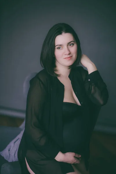 Young pregnant woman with dark hair in black lingerie and a black silk shirt on a dark background