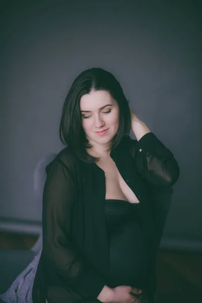 Young pregnant woman with dark hair in black lingerie and a black silk shirt on a dark background