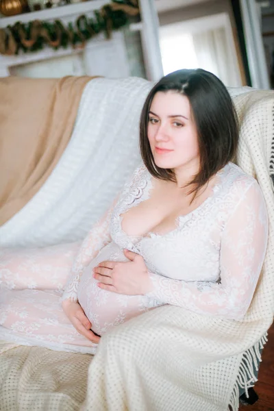 クリスマスの設定でソファの上にレースの白いドレスで黒髪の若い妊娠中の女の子 — ストック写真