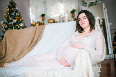 Bir Noel ortamında kanepeüzerinde dantel beyaz elbise koyu saçlı genç hamile kız