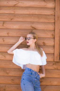 Mükemmel vücutlu çekici bir genç kız ve son moda kotlu sarı saçlar ve yazın ahşap bir duvara karşı fırfırlı ve güneş gözlüklü beyaz bir bluz.