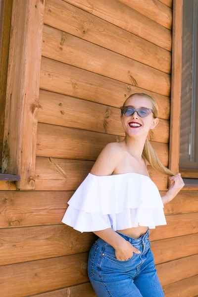 Attractive young girl with a perfect figure and blond hair in trendy jeans and a white top with ruffles and sunglasses in the summer against a wall of wood