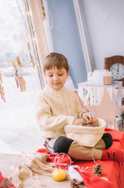 Sıcak beyaz örülü süveter giymiş şirin bir çocuk Noel için pencerenin kenarında oturur. Battaniyeler, koniler, mandalinalar ve zencefilli kurabiyelerle süslenir. Noel havası