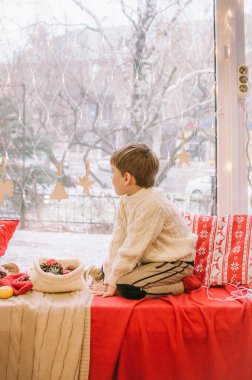 Sıcak beyaz örülü süveter giymiş şirin bir çocuk Noel için pencerenin kenarında oturur. Battaniyeler, koniler, mandalinalar ve zencefilli kurabiyelerle süslenir. Noel havası