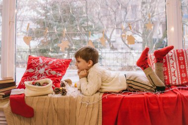 Sıcak beyaz örülü süveter giymiş şirin bir çocuk Noel için pencerenin kenarında oturur. Battaniyeler, koniler, mandalinalar ve zencefilli kurabiyelerle süslenir. Noel havası
