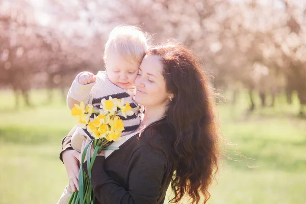 Family in spring park with spring flowers. A young mother with long dark hair with a small son cuddles with her baby son in spring. Spring mood. Mothers Day