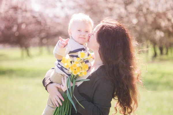 春の公園で春の花と家族 幼い息子と長い黒い髪をした若い母親が春に赤ん坊の息子と抱き合う 春の気分 母の日 — ストック写真