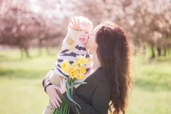 Family in spring park with spring flowers. A young mother with long dark hair with a small son cuddles with her baby son in spring. Spring mood. Mothers Day