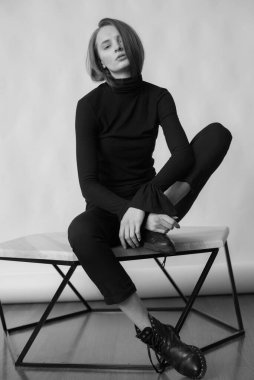 Sarı saçlı, siyah diz çoraplı, siyah pantolonlu, beyaz arka planda, stüdyoda poz veren, çıplak makyajlı genç ve çekici bir manken.