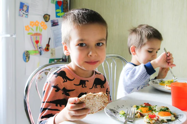 Çocuklar mutfakta masada oturuyorlar ve çırpılmış yumurta ve sebzelerle kahvaltı yapıyorlar.