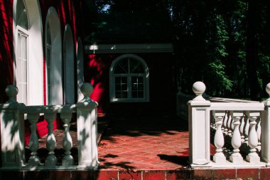 Kemerli yarı dairesel pencereli, beyaz balkon korkulukları olan, ağaçların arasına gömülü, rahat, beyaz ve kırmızı bir ev.