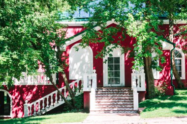 Kemerli yarı dairesel pencereli, beyaz balkon korkulukları olan, ağaçların arasına gömülü, rahat, beyaz ve kırmızı bir ev.