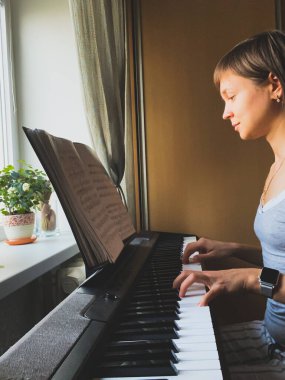 Açık gri bluzlu genç bir kız bir sandalyeye oturur ve elektronik piyano çalar, önünde notalar vardır.