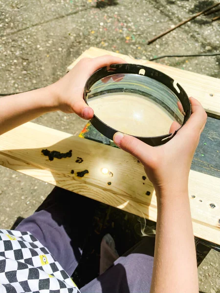 Bir çocuk sokakta oturur ve bir tahta parçasını yakmak için büyüteç kullanır.