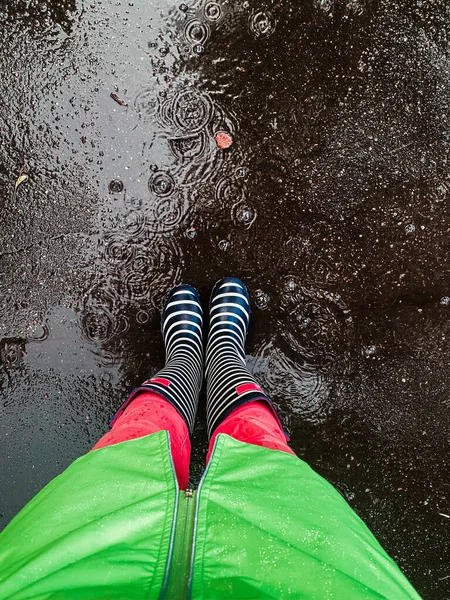 Çizgili lastik çizmeli ve yeşil yağmurluklu bir kız ıslak bir kaldırımda duruyor, bacaklarının üst görüntüsü.