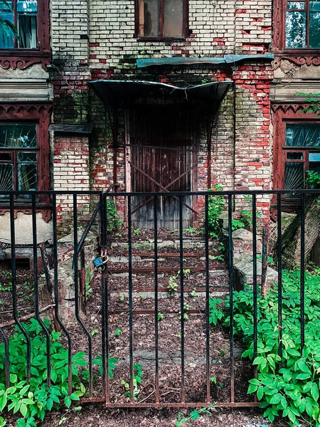 Paslı bir çitin arkasında terk edilmiş tuğla bir ev. Çimlerle kaplı harap basamaklar.