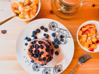 Tahta bir masada, krep, yaban mersini, meyve kasesi ve bir çaydanlık yeşil çay.