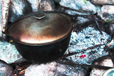 Tütsülenmiş demir bir tencere taştan yapılmış bir şöminenin içinde bir ormanın arka planında duruyor.