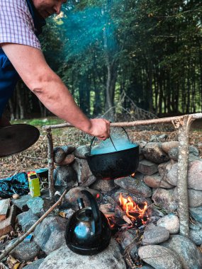 Bir adam tencerede yemek hazırlar ve kepçeyle karıştırır, yanında da tütsülenmiş teneke çaydanlık vardır.