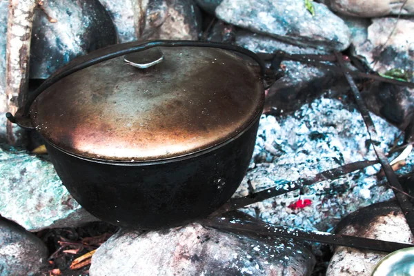 Tütsülenmiş demir bir tencere taştan yapılmış bir şöminenin içinde bir ormanın arka planında duruyor.