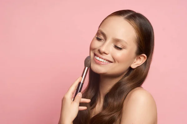 Kecantikan makeup. Gadis muda dengan kuas makeup di tangan pada latar belakang merah muda — Stok Foto