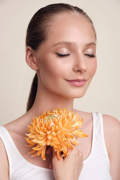 Gesichtsschönheit. Blumen und ein Modell-Porträt. Mädchen, das eine Blume mit geschlossenen Augen vor beigem Hintergrund in die Kamera hält. Mädchen mit natürlichem Nackt-Make-up und gesunder Haut — Stockfoto